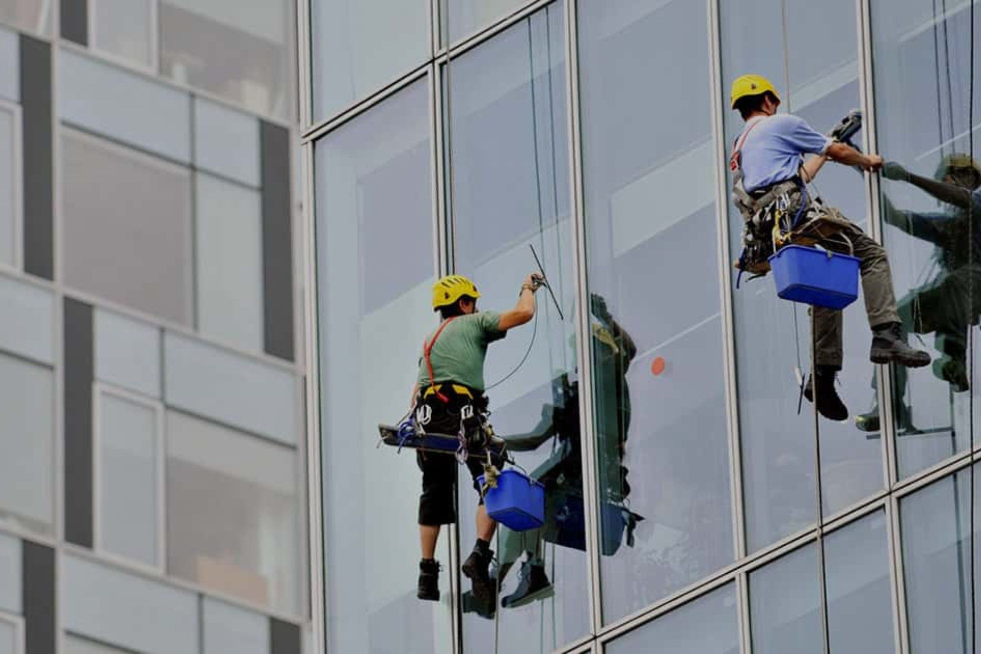 Dos personas con cascos amarillos y arneses, suspendidas mediante cuerdas frente a una fachada acristalada, con utensilios y cubos azules.